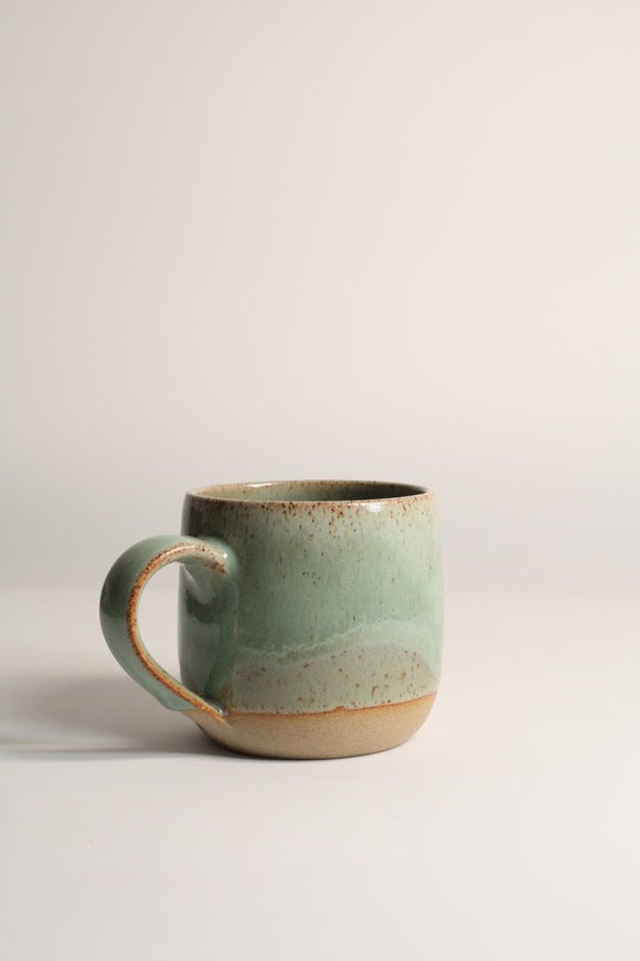 A handcrafted ceramic mug with a beige base color and green speckled patterns, displayed against a neutral background.