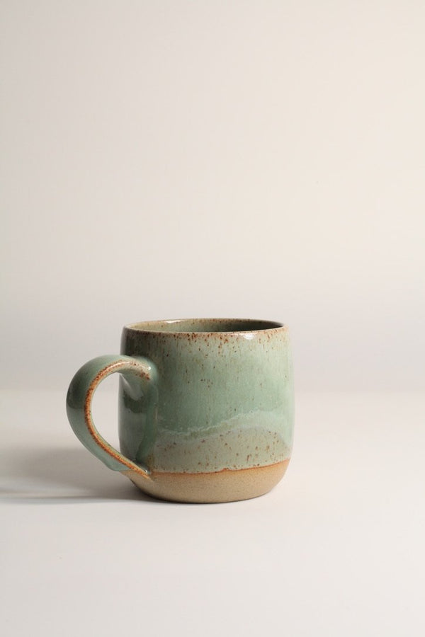 A handcrafted ceramic mug with a beige base color and green speckled patterns, displayed against a neutral background.