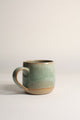 A handcrafted ceramic mug with a beige base color and green speckled patterns, displayed against a neutral background.