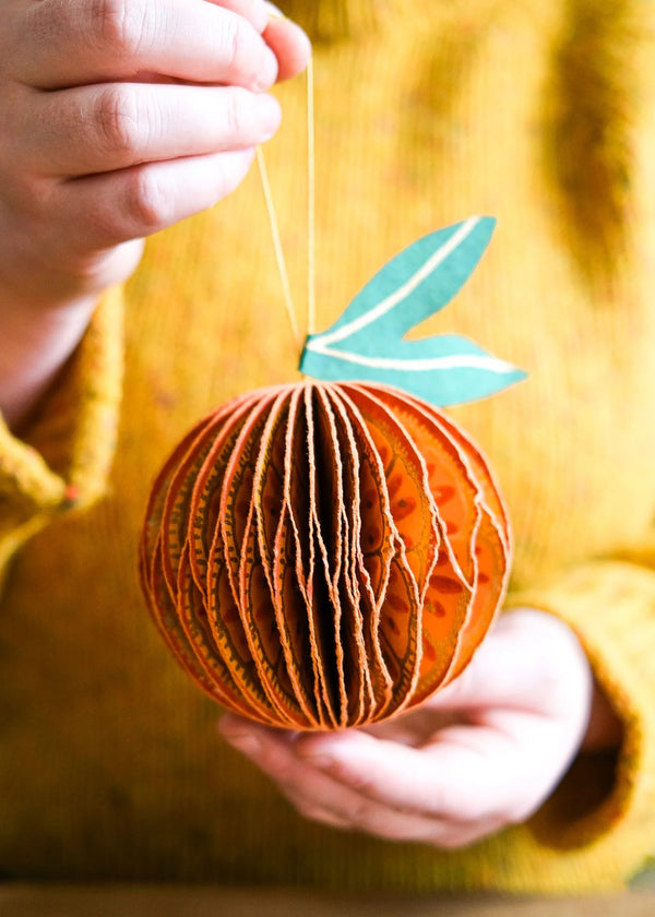 Hand holding a decorative paper ornament shaped like a pumpkin with a green leaf against a blurred yellow background.