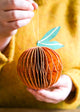 Hand holding a decorative paper ornament shaped like a pumpkin with a green leaf against a blurred yellow background.