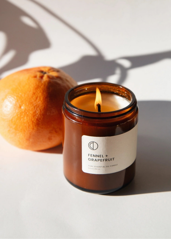 Candle in an amber jar with a label next to an orange on a light background