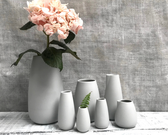 A collection of porcelain vases in subtle colors, with a larger vase in the center containing a pink flower, and four smaller vases in front of it.