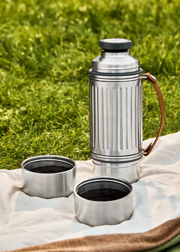 Silver thermos and two cups on a blanket with grass in the background