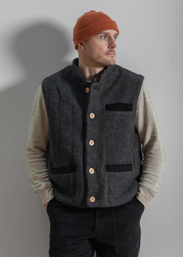 Man wearing a gray vest over a beige sweater with a red beanie against a plain background