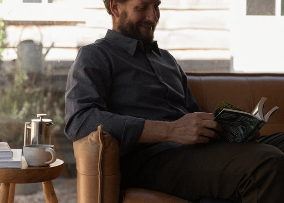 Man sitting on a brown leather couch reading a book in a cozy room.