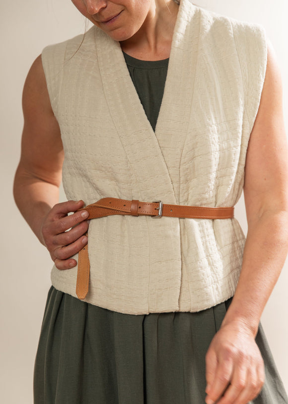 Person wearing a beige vest with a brown belt and green skirt against a neutral background