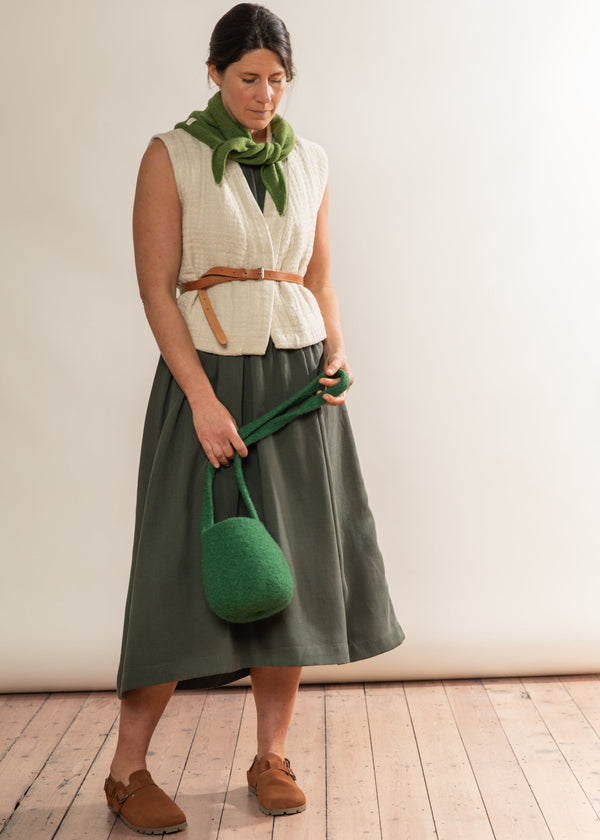 Woman in a white sleeveless top and green skirt holding a green bag against a plain background