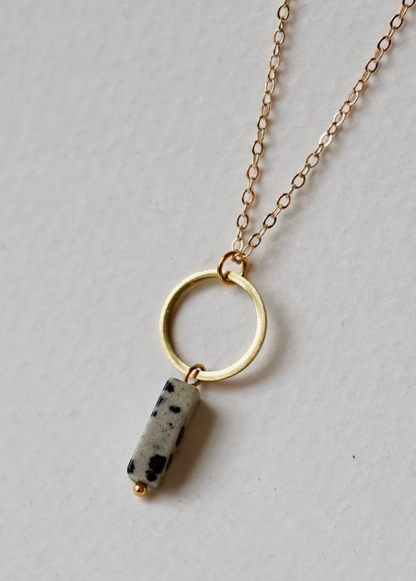 A Dalmatian Jasper and Gold Circle Necklace laid out on a plain surface, featuring a gold-plated hoop with a unique bead of dalmatian jasper on a delicate chain.