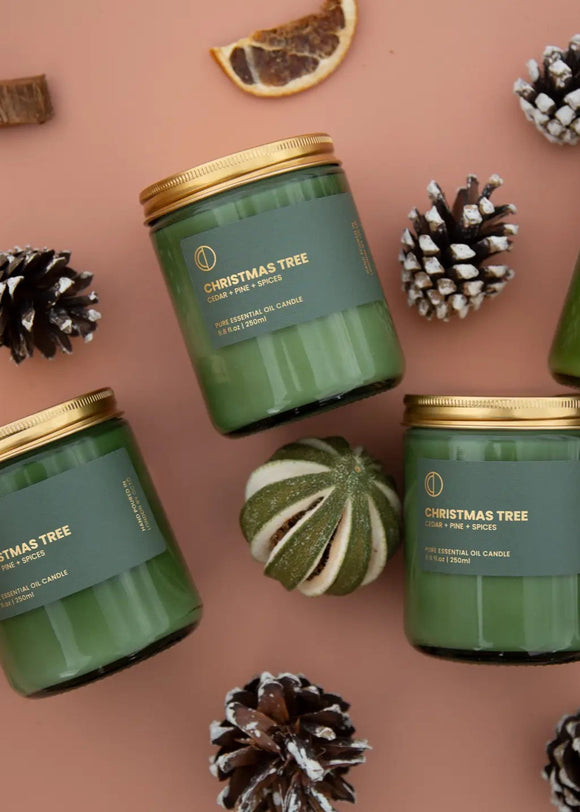 Multiple green jar candles with gold lids, labeled 'Merry Christmas', displayed among pine cones and citrus fruit decorations.