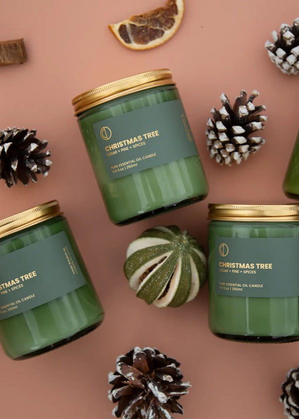 Multiple green jar candles with gold lids, labeled 'Merry Christmas', displayed among pine cones and citrus fruit decorations.