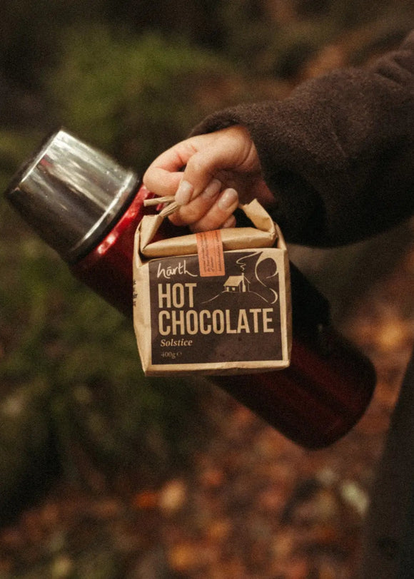 A person holding a red thermos with a brown package labeled 'HARTH HOT CHOCOLATE' in a natural outdoor setting.
