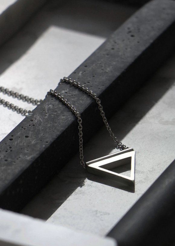 A sterling silver necklace with a geometric triangle symbol on a chain, displayed against a concrete and marble background.