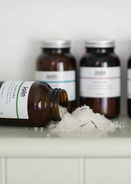 Four bottles of luxury bath salts on a shelf with the front bottle倾倒着, spills salts. The labels read 'Joan', 'John', 'Jane', and 'Bob'.