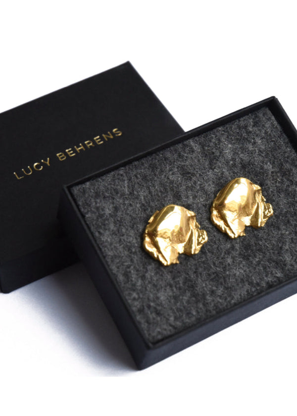 A pair of gold-colored earrings displayed in an open black case with 'LUCY BEHRENS' written on it, set on a gray velvet lining.