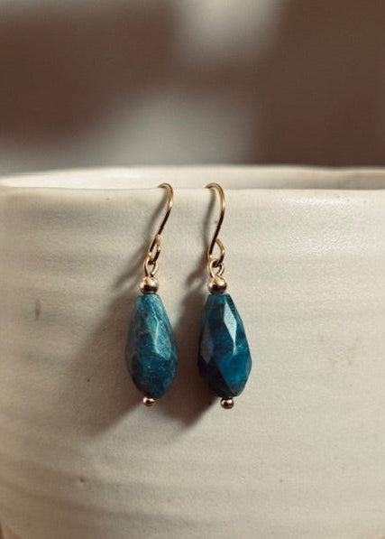 Teal drop earrings with gold hooks on a white ceramic surface