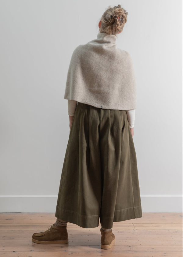 Person wearing a beige turtleneck and olive green wide-leg pants standing against a white wall.