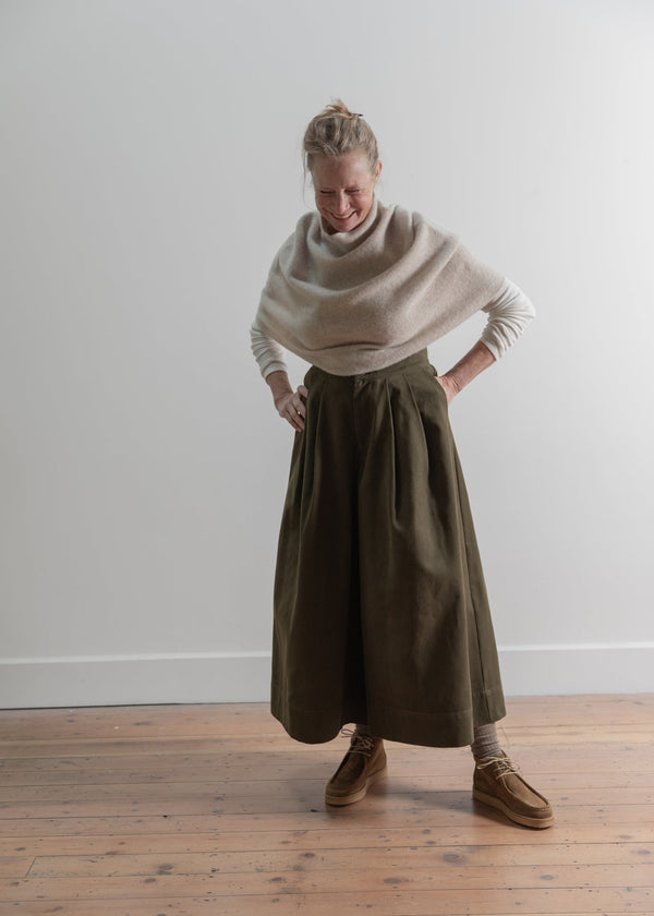 Person wearing a beige sweater and olive green skirt standing on a wooden floor against a white wall.