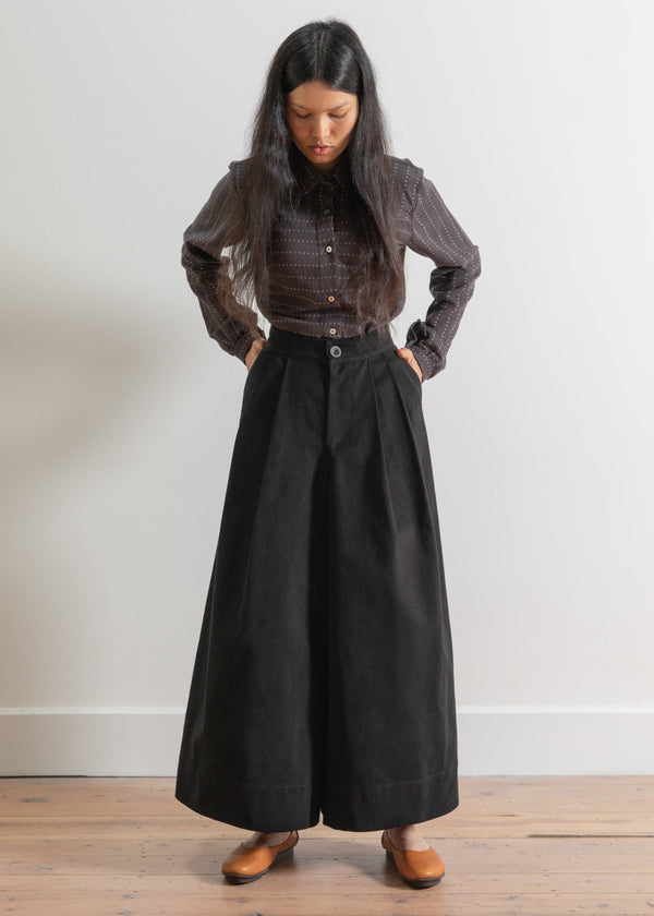Woman wearing a dark outfit with a long skirt and blouse against a white wall.