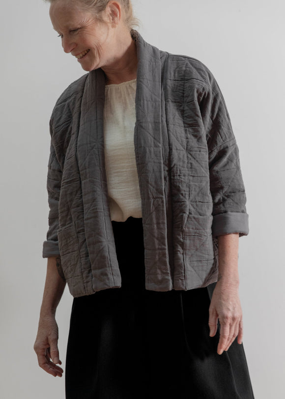 Woman wearing a gray quilted jacket over a white top and black skirt on a plain background