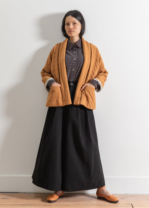 Person wearing a mustard coat and black skirt against a white wall