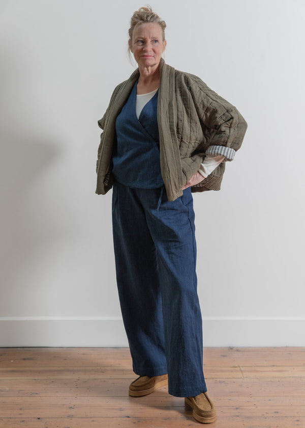 Woman wearing a green jacket and blue pants standing against a white wall.