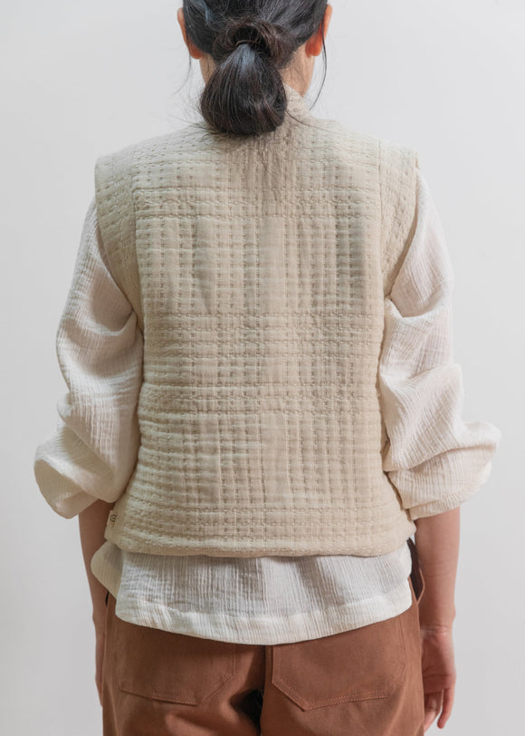 Person wearing a beige quilted vest over a white shirt with brown pants on a plain background