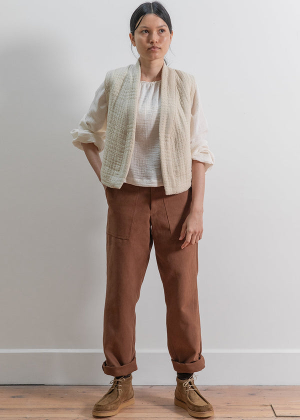 Woman wearing a beige jacket, white shirt, brown pants, and brown shoes against a plain background