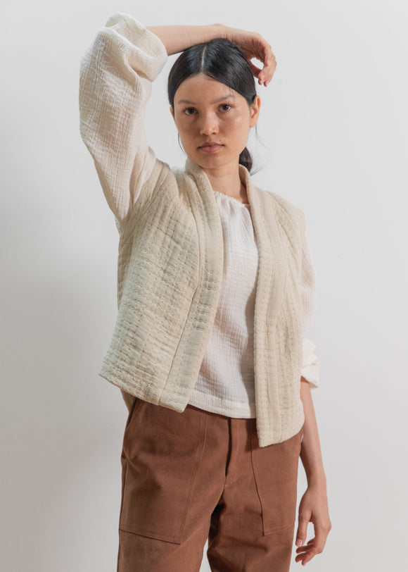 Woman wearing a beige cardigan over a white shirt and brown pants on a plain background