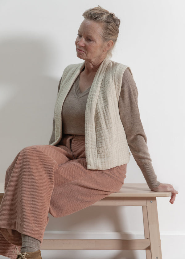 Woman sitting on a wooden stool wearing a beige cardigan and brown pants against a white background