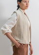 Woman wearing a beige textured vest over a white shirt with brown pants on a plain background