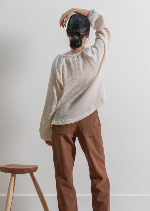 Person wearing a beige sweater and brown pants standing against a white wall with a wooden stool.