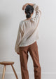 Person wearing a beige sweater and brown pants standing against a white wall with a wooden stool.