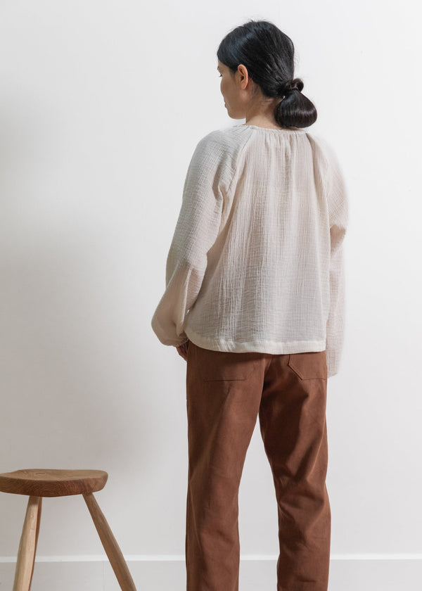 Person wearing a light-colored blouse and brown pants standing against a white wall with a wooden stool.