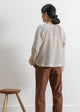Person wearing a light-colored blouse and brown pants standing against a white wall with a wooden stool.