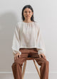 Woman sitting on a stool wearing a white blouse and brown pants against a plain background