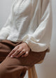 Person wearing a white textured sweater and brown pants sitting on a stool.