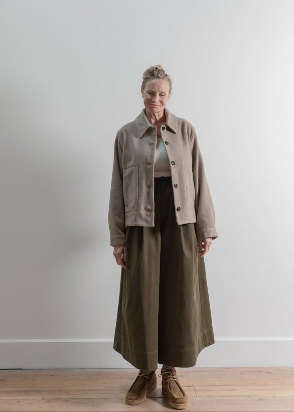 Person wearing a beige jacket and long brown coat against a white wall.