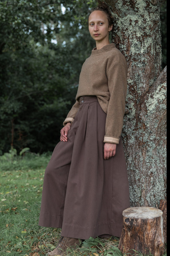 A woman stands outdoors leaning against a tree stump, wearing a brown knit sweater with light beige cuffs and a long brown very wide leg trousers. The background shows a grassy forest setting.