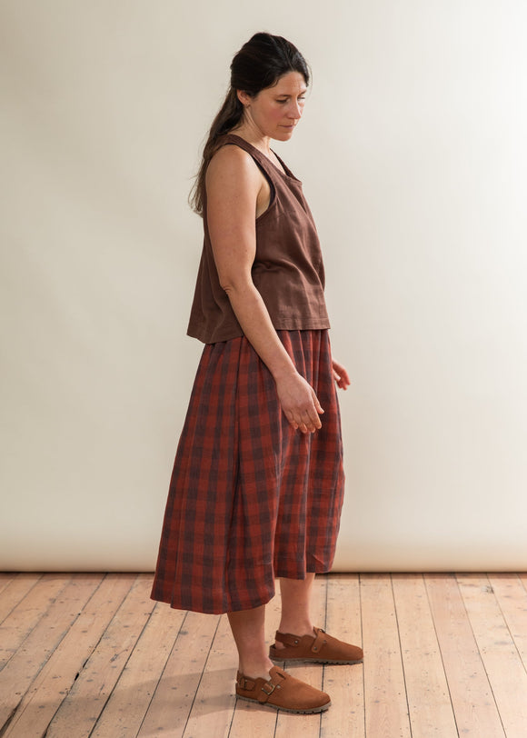 Woman wearing a brown sleeveless top and plaid skirt standing on a wooden floor against a white wall.