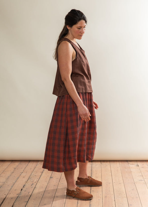 Woman wearing a brown sleeveless top and plaid skirt standing on a wooden floor against a white wall.