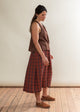 Woman wearing a brown sleeveless top and plaid skirt standing on a wooden floor against a white wall.