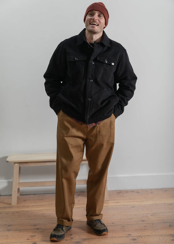 Man wearing a black jacket and brown pants standing in a room with a wooden floor and white wall.
