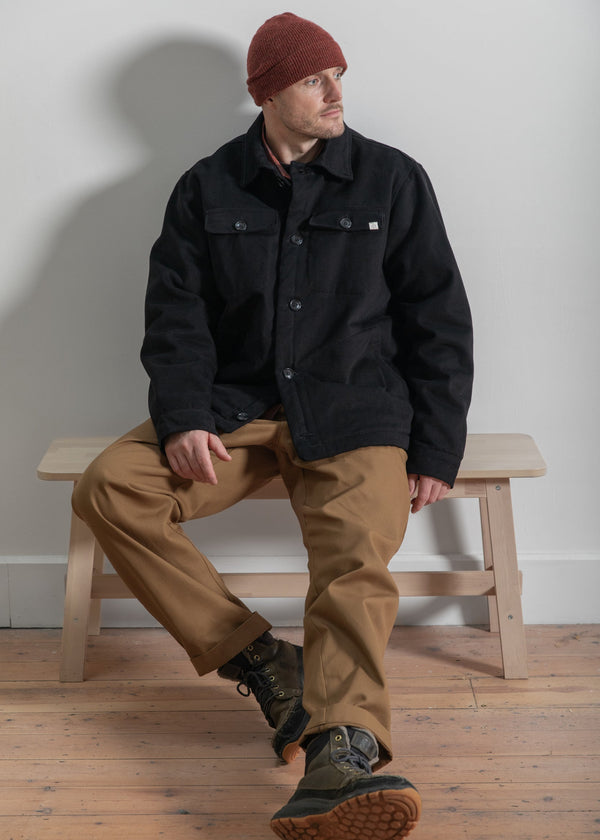Man sitting on a wooden bench wearing a black jacket, brown pants, and a red beanie.
