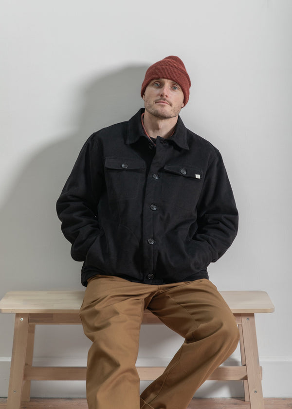 Man wearing a black jacket and brown pants sitting on a wooden bench against a plain wall.