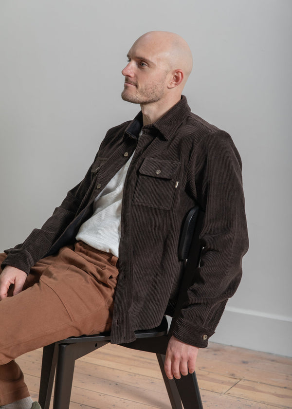 Man sitting on a chair wearing a brown jacket and brown pants against a plain background