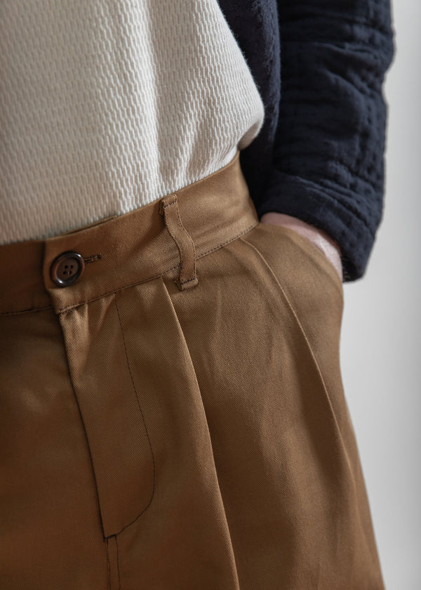 Close-up of brown pants with a button, worn by a person, on a plain background