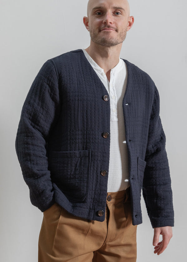 Man wearing a navy cardigan over a white shirt with brown pants on a plain background