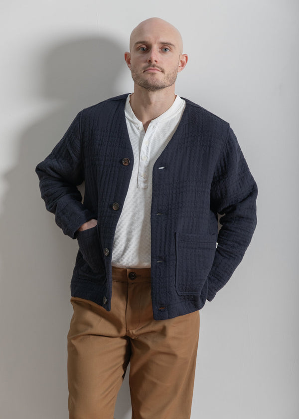 Man wearing a navy cardigan over a white shirt and brown pants against a plain background