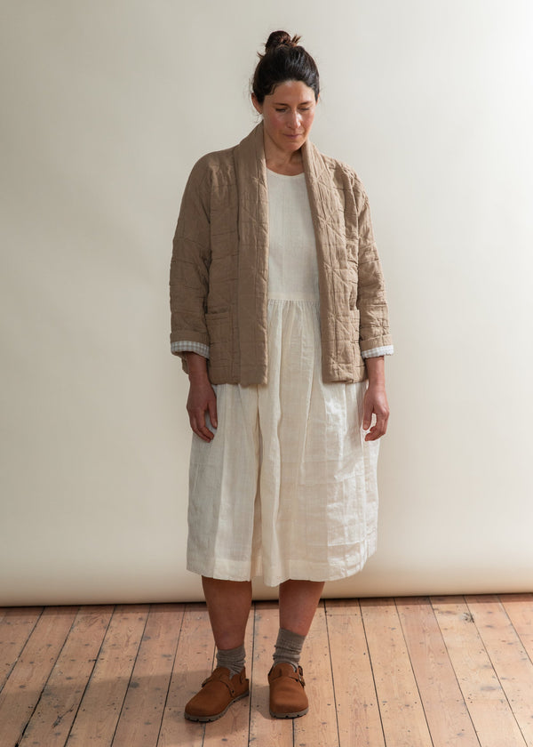 Woman wearing a beige cardigan, white dress, and brown shoes in a room with wooden flooring.